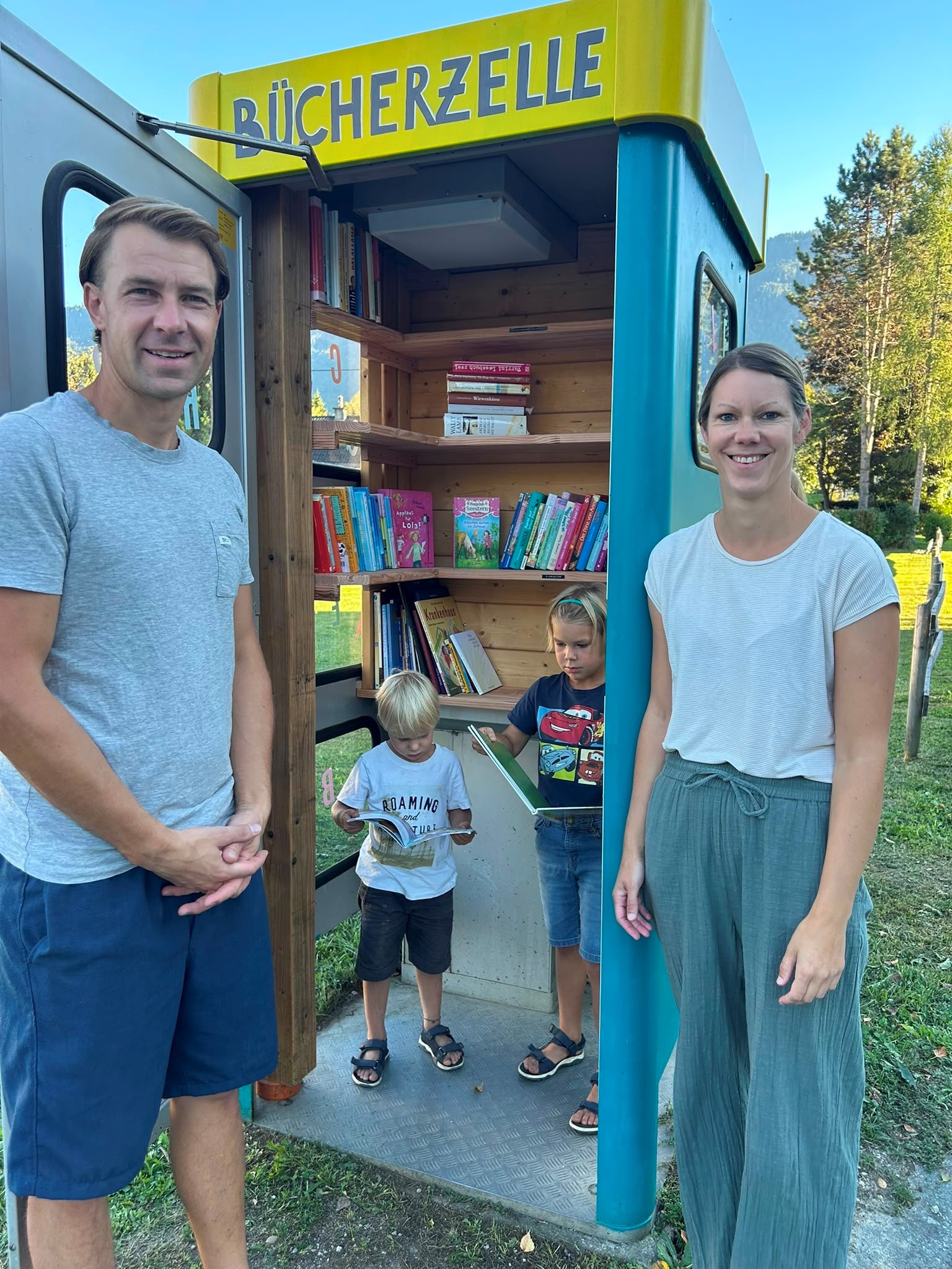 Familie vor Büchertauschzelle in Tirol mit Kindern beim Lesen von Büchern.